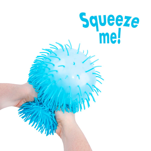 A bright blue squeeze ball stress toy with spiky protrusions, being squeezed by two hands against a white background, with the text 'Squeeze me!' in blue in the upper right.