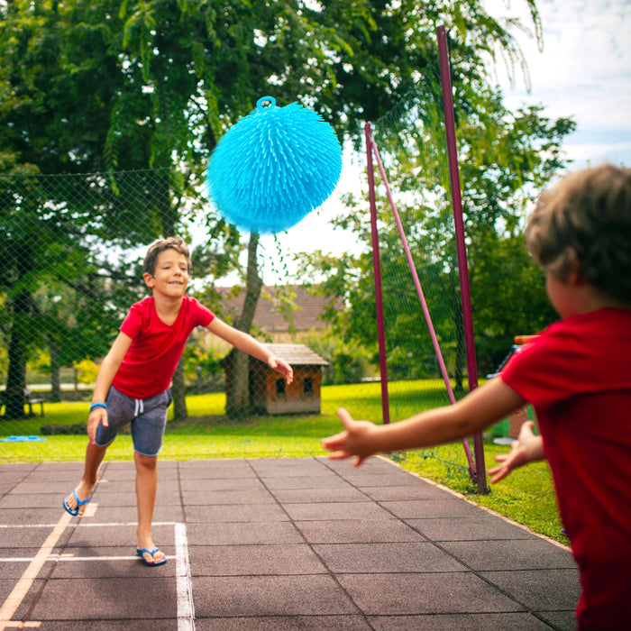 Two children playing with a bright blue textured hanging toy outdoors on a paved backyard court, with trees and a fence in the background.
