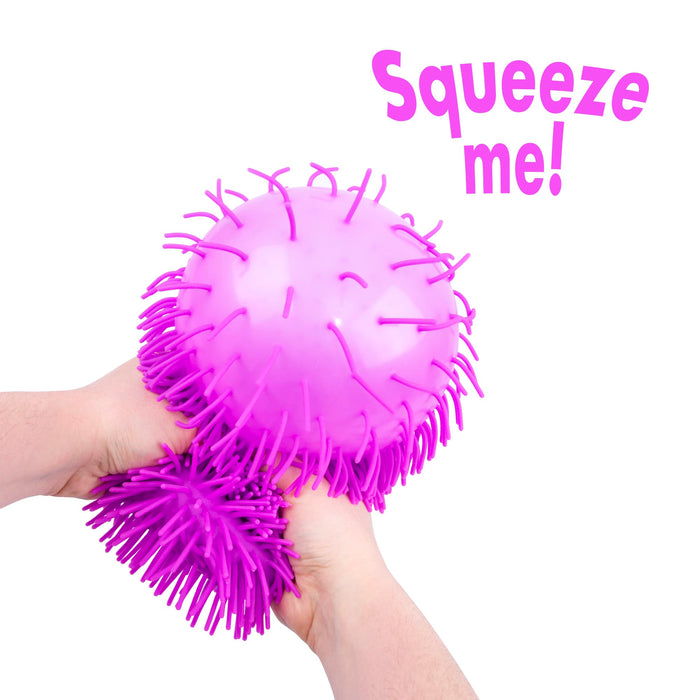 A pair of hands holding a bright pink squeeze ball toy with long spines, with a matching pink spiky textured sleeve resting underneath. The text 'Squeeze me!' is written in pink in the upper right corner on a solid white background.
