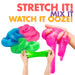 Three hands stretching colorful slime against a white background with bold text reading 'STRETCH IT! WATCH IT OOZE!' and 'MIX IT'. The slime colors include bright pink, blue, green, and a stretched green section.