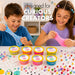 Two children smiling and stretching colorful slime at a table with multiple small slime containers labeled 'Curious Creators' and scattered beads and craft pieces. Large text at the top reads 'designed for CURIOUS CREATORS'.