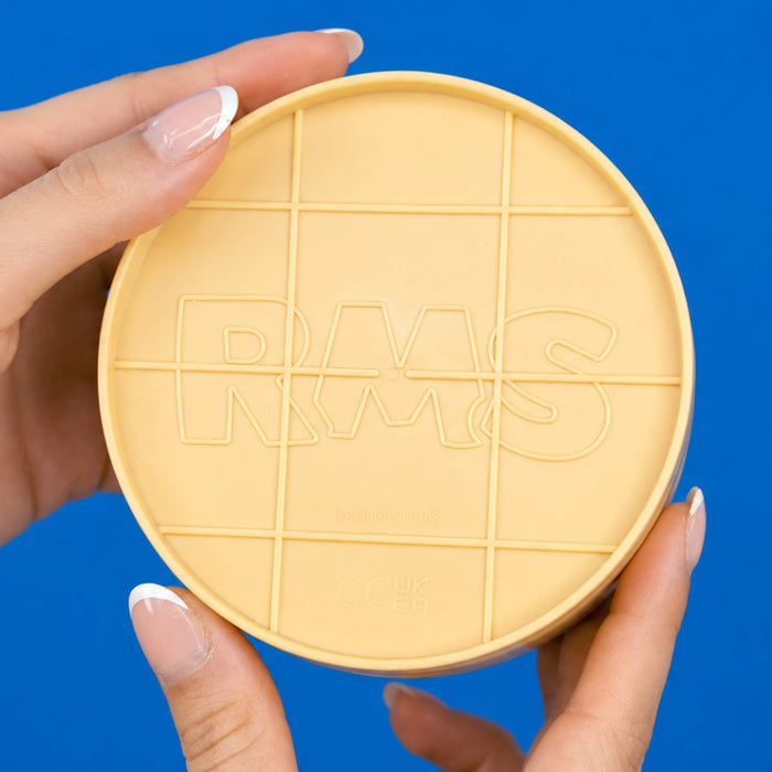 A person's hands holding a round, pale yellow cosmetic container lid with a grid pattern and the embossed letters 'RMS' against a solid blue background.