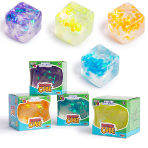 Four colourful translucent stress cubes shown with their retail boxes on a white background. The cubes come in assorted pastel and bright colors, including purple, yellow-green, peach, blue, and teal, with some containing glitter or sparkly inclusions.