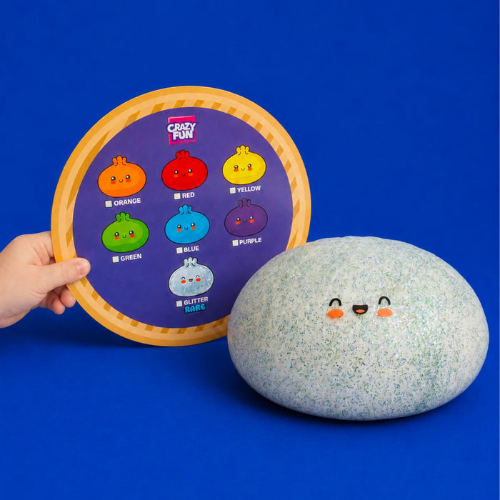 A hand holds a round purple toy board labeled 'Crazy Fun' with colorful cartoon plop-plop characters and names, next to a large speckled gray-green squishy ball with a smiling face, on a solid blue background. Which colour will you receive?