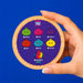 A hand holds a round wooden box lid featuring a purple insert with colorful cartoon pomegranate illustrations labeled Green, Pink, Purple, Red, Blue, Yellow, and Rainbow Rare, against a solid blue background.