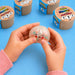 A person's hands holding a clear, bubble-shaped squishy toy filled with multicolor glitter and a cute face, with several colorful 'Squishy World' mystery rolls in the background on a blue surface.