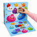 A colourful advent-style gift box with numbered cartoon dumpling faces on a light blue box, shown open with a flat lower panel. A hand in a pink sleeve is holding a small wrapped gift. Contains 24 mini squishy dumplings.