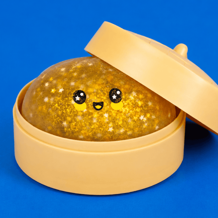 A round glittery gold quackling stress ball with a cute face, sitting inside a beige plastic container with its lid propped open, set against a solid blue background.