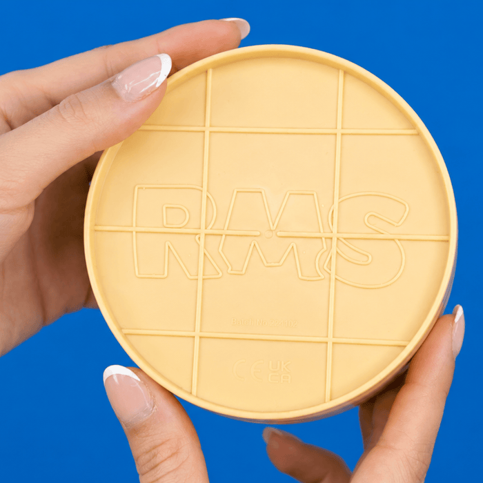 A person's hands holding a round, light yellow cosmetic compact lid with a grid pattern and the embossed letters 'RMS' against a solid blue background. Shows authenticity with the RMS logo on the bottom. 