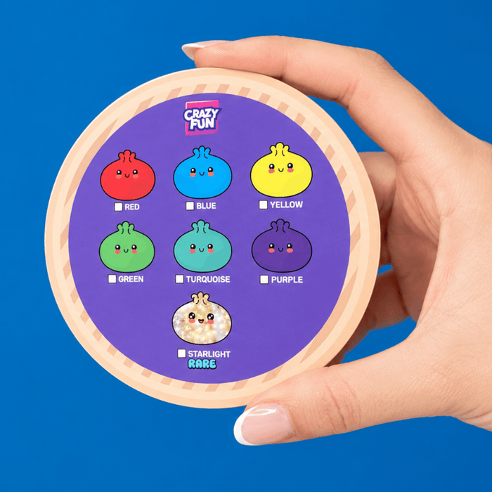 A hand holding a round cosmetic compact against a solid blue background. The compact lid features a purple center with cartoon pomegranate-style color options labeled Red, Blue, Yellow, Green, Turquoise, Purple, and Starlight Rare, under the brand name Crazy Fun.
