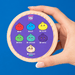 A hand holding a round cosmetic compact against a solid blue background. The compact lid features a purple center with cartoon pomegranate-style color options labeled Red, Blue, Yellow, Green, Turquoise, Purple, and Starlight Rare, under the brand name Crazy Fun.